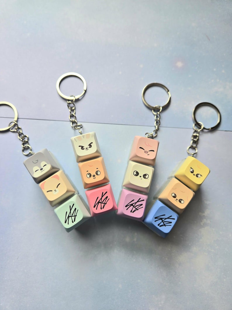 May include: Keyrings with stacked, colourful keycaps featuring cartoon animal faces and abstract designs. Each keyring has a silver ring and chain. The keycaps are in shades of grey, peach, pink, yellow, and blue.