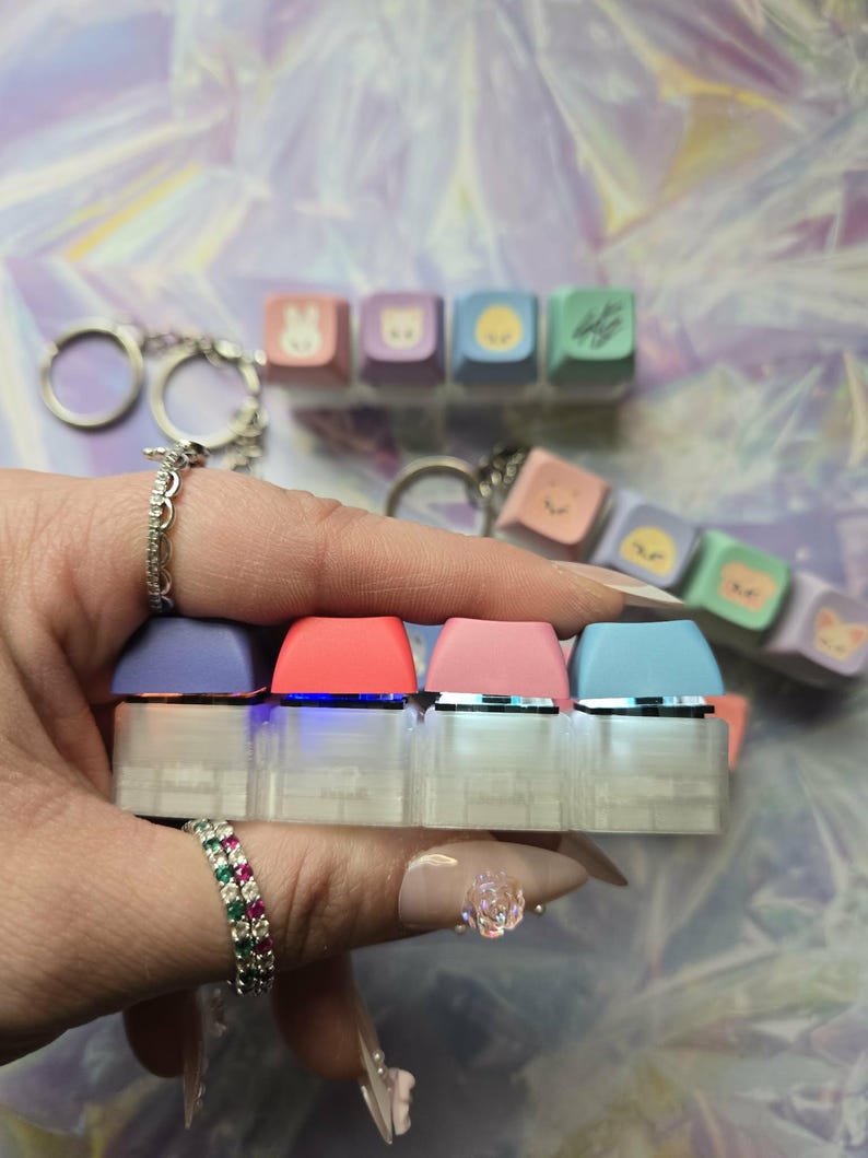 May include: A miniature keyboard with four keycaps in purple, red, pink, and blue. Additional keycaps with cartoon faces are attached to a keyring. The keyboard is held in a hand. The keyboard is clear.