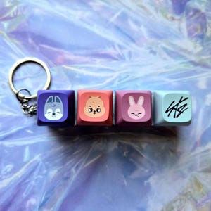 May include: A keyring with four square keycaps in various colours. Each keycap displays a different design: a purple keycap with a cartoon wolf, a coral keycap with a cartoon hamster, a pink keycap with a cartoon rabbit, and a light blue keycap with a stylized design.