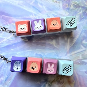 May include: Keychains with colourful keyboard keycaps, each with a cute animal design. The keychains have five keycaps in shades of pink, purple, and orange. They are attached to a silver chain and keyring.