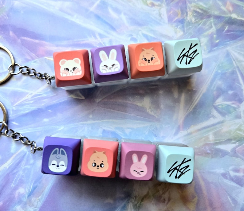 May include: Keychains with colourful keyboard keycaps, each with a cute animal design. The keychains have five keycaps in shades of pink, purple, and orange. They are attached to a silver chain and keyring.