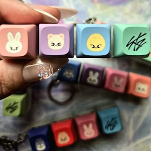 May include: A selection of colourful keycaps with cartoon animal designs and abstract symbols. The keycaps are in shades of pink, purple, blue, and green. Some keycaps are attached to keychains. The image is well-lit and the keycaps are in focus.