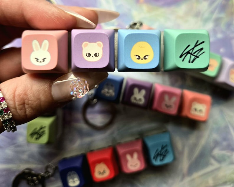 May include: A selection of colourful keycaps with cartoon animal designs and abstract symbols. The keycaps are in shades of pink, purple, blue, and green. Some keycaps are attached to keychains. The image is well-lit and the keycaps are in focus.