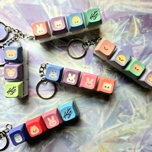 May include: Keyrings with colourful square charms, each displaying a cartoon animal face and stylized text. The charms are attached to a silver chain and keyring. The charms are in pastel shades of pink, blue, purple, and green.