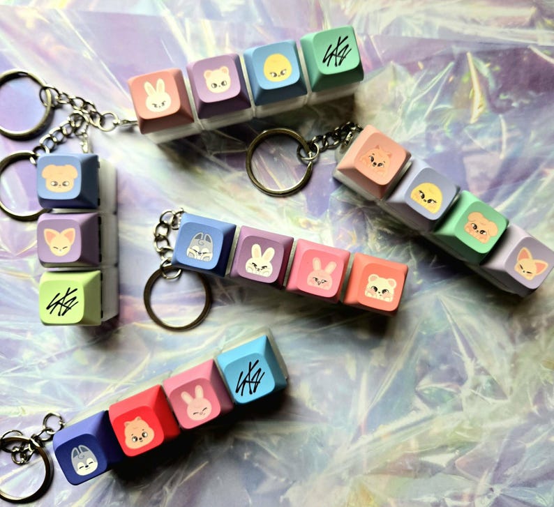 May include: Keyrings with colourful square charms, each displaying a cartoon animal face and stylized text. The charms are attached to a silver chain and keyring. The charms are in pastel shades of pink, blue, purple, and green.