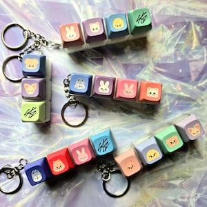 May include: Keyrings with colourful square keycaps, each decorated with cartoon animal faces and symbols. The keyrings have silver chains and round key rings. The keycaps are in shades of blue, pink, purple, and green.