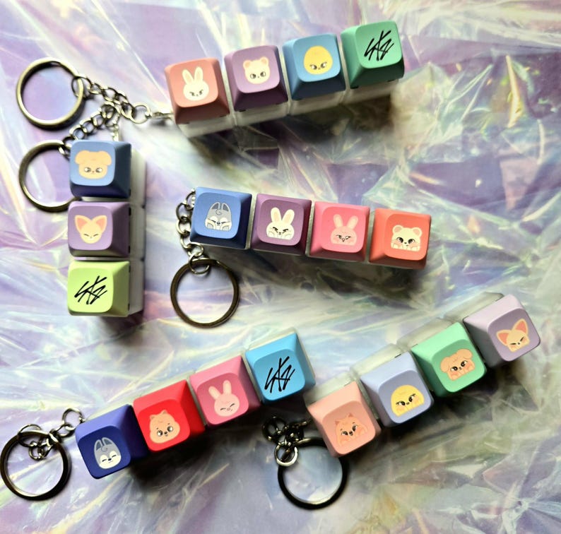 May include: Keyrings with colourful square keycaps, each decorated with cartoon animal faces and symbols. The keyrings have silver chains and round key rings. The keycaps are in shades of blue, pink, purple, and green.