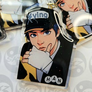 May include: Clear acrylic keychain featuring an anime-style illustration of a person wearing a black cap with the word "Xying" and the name "HAN". The person has blue hair and is wearing a black suit with a white glove. The keychain has a gold-colored ring.