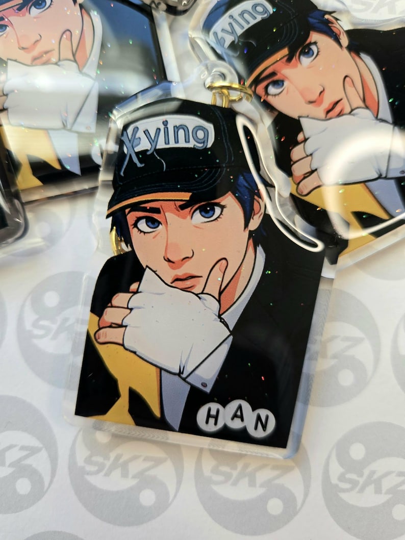 May include: Clear acrylic keychain featuring an anime-style illustration of a person wearing a black cap with the word "Xying" and the name "HAN". The person has blue hair and is wearing a black suit with a white glove. The keychain has a gold-colored ring.