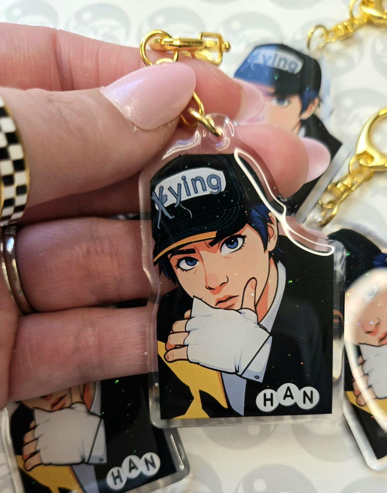 May include: Clear acrylic keychain featuring a stylized anime-style illustration of a person with blue hair and eyes, wearing a black cap with the word "Xying". The keychain has a gold-colored clasp and the name "HAN" is printed on the bottom.
