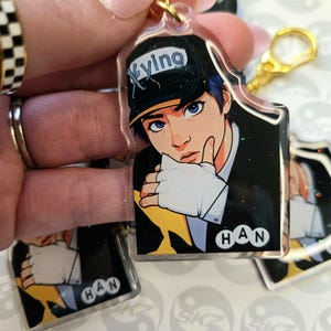 May include: Clear acrylic keychain featuring a stylized illustration of a person with blue hair and eyes, wearing a black cap with the word "Xyving". The keychain has a gold-colored clasp and the name "HAN" is printed on the bottom.