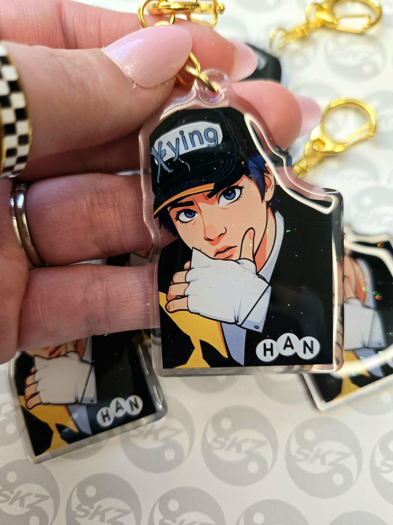 May include: Clear acrylic keychain featuring a stylized illustration of a person with blue hair and eyes, wearing a black cap with the word "Xyving". The keychain has a gold-colored clasp and the name "HAN" is printed on the bottom.