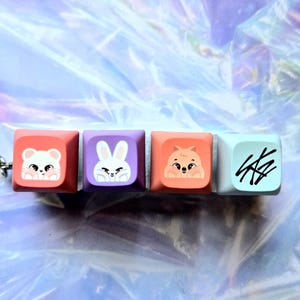 May include: A keychain with four square keycaps, each with a different cartoon animal design. The keycaps are in pink, purple, orange, and light blue. The first keycap has a white mouse, the second a white rabbit, the third a brown hamster, and the fourth has black scribbles.