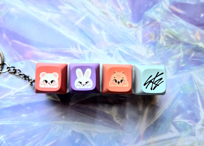 May include: A keychain with four square keycaps, each with a different cartoon animal design. The keycaps are in pink, purple, orange, and light blue. The first keycap has a white mouse, the second a white rabbit, the third a brown hamster, and the fourth has black scribbles.
