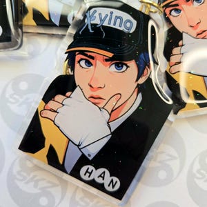 May include: A clear acrylic keychain featuring a stylized illustration of a person with blue hair and eyes, wearing a black cap with the word "Xying". The person is wearing a black suit jacket and white gloves. The name "HAN" is printed at the bottom.