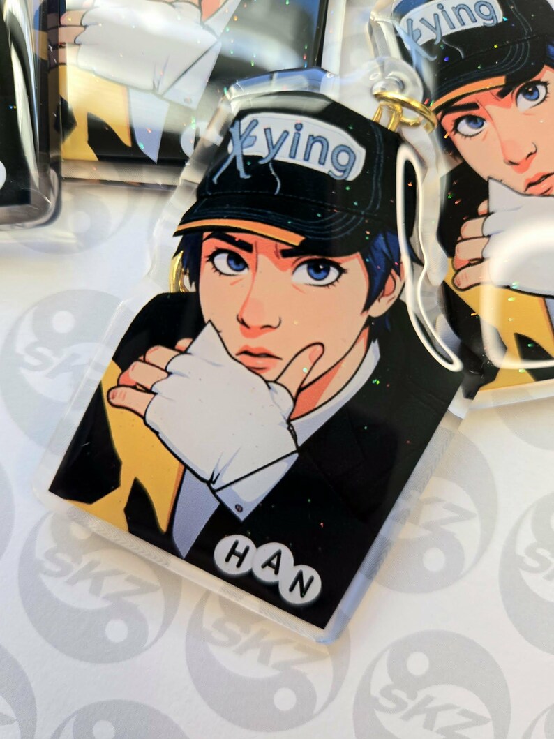 May include: A clear acrylic keychain featuring a stylized illustration of a person with blue hair and eyes, wearing a black cap with the word "Xying". The person is wearing a black suit jacket and white gloves. The name "HAN" is printed at the bottom.