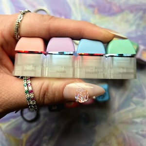 May include: A close-up of four pastel-coloured keyboard keycaps. The keycaps are pink, lilac, light blue, and mint green. They are held together by a clear, rectangular base. Each keycap has a silver trim and is lit from below with a red light.