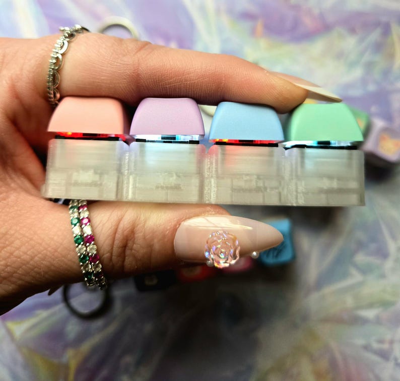May include: A close-up of four pastel-coloured keyboard keycaps. The keycaps are pink, lilac, light blue, and mint green. They are held together by a clear, rectangular base. Each keycap has a silver trim and is lit from below with a red light.