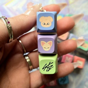 May include: A stack of three colourful square charms. The top charm is light blue with a cartoon dog face, the middle is purple with a fox face, and the bottom is green with black text. A silver chain is attached to the top charm.