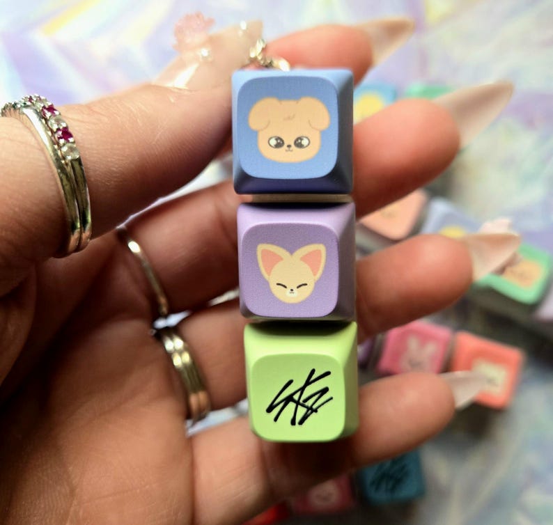 May include: A stack of three colourful square charms. The top charm is light blue with a cartoon dog face, the middle is purple with a fox face, and the bottom is green with black text. A silver chain is attached to the top charm.