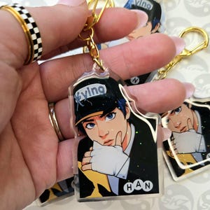 May include: Clear acrylic keychain featuring a stylized illustration of a person with blue hair and eyes, wearing a black cap with the word "Xyung". The keychain has a gold-colored clasp and a gold-colored ring. The name "HAN" is printed on the keychain.