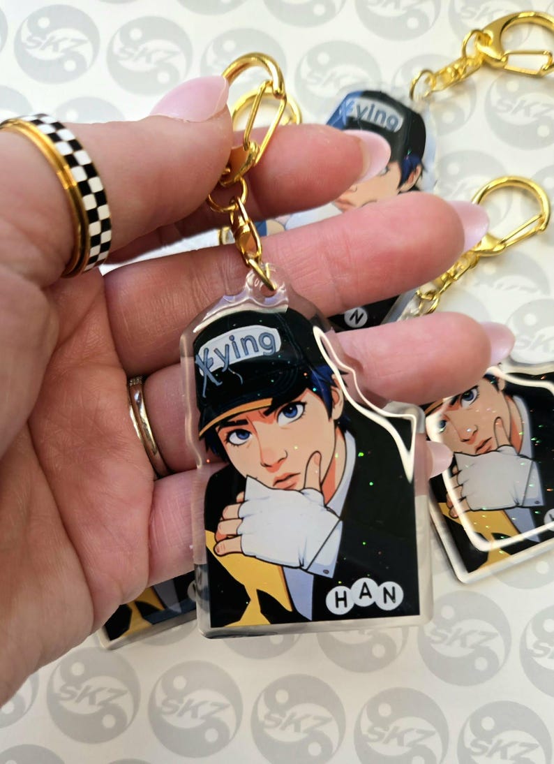 May include: Clear acrylic keychain featuring a stylized illustration of a person with blue hair and eyes, wearing a black cap with the word "Xyung". The keychain has a gold-colored clasp and a gold-colored ring. The name "HAN" is printed on the keychain.