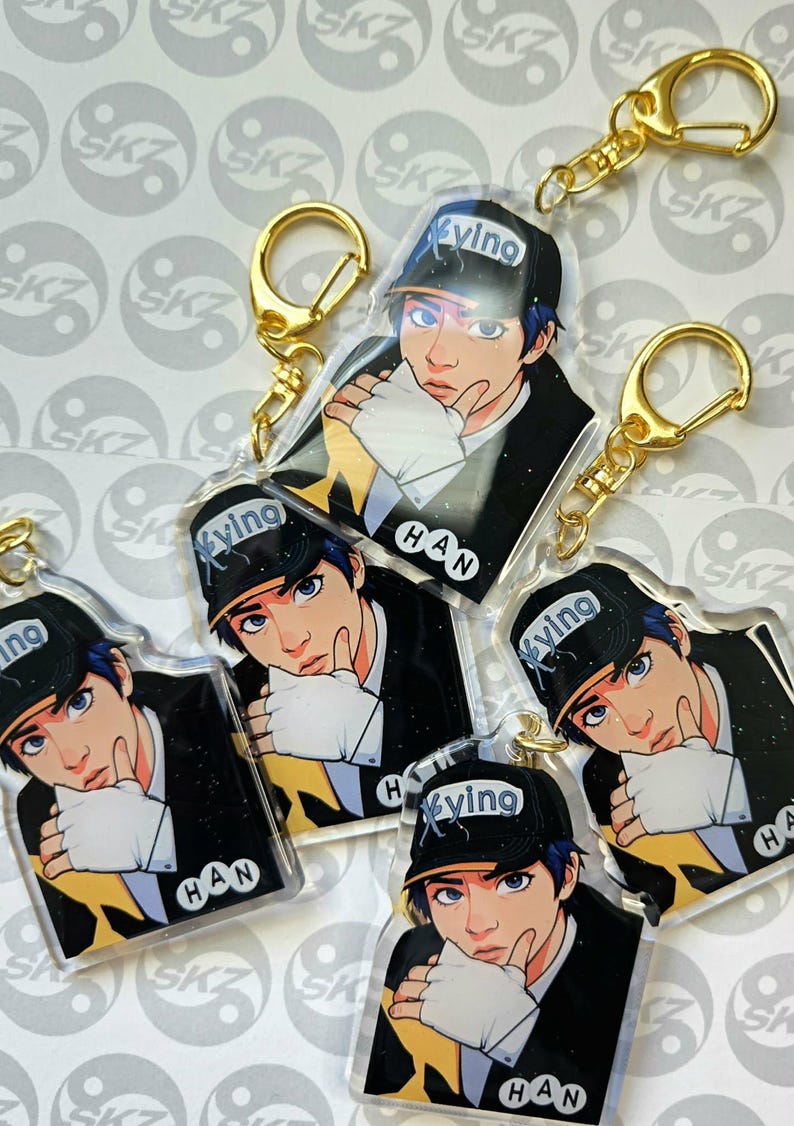 May include: A collection of clear acrylic keychains featuring a stylized illustration of a person wearing a black cap with the word "ying" and the name "HAN". Each keychain has a gold-colored clasp.