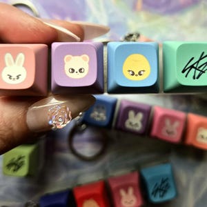 May include: A set of five colourful keycaps featuring cartoon animal and symbol designs. The keycaps are pink, purple, blue, and green. The keycaps are held by a hand. Additional keycaps are visible in the background.