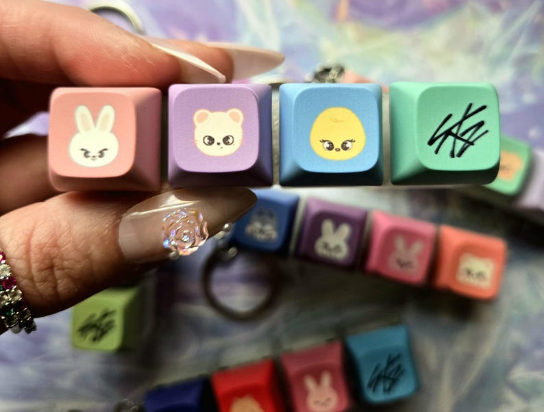 May include: A set of five colourful keycaps featuring cartoon animal and symbol designs. The keycaps are pink, purple, blue, and green. The keycaps are held by a hand. Additional keycaps are visible in the background.