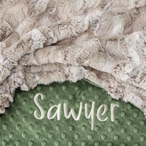 May include: A soft, beige faux fur blanket folded over a sage green minky fabric backing with a raised dot texture. The name Sawyer is embroidered in white cursive script across the centre of the green fabric section of the blanket.