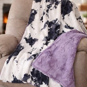 May include: A plush blanket with a cow-print design in black and white, draped over a beige recliner. The blanket has a soft, textured appearance. A solid lavender blanket is folded over the top.