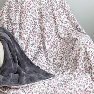 May include: A soft, plush throw blanket with a pink and gray leopard print on one side and a solid gray plush on the other side. The blanket is draped over a chair.