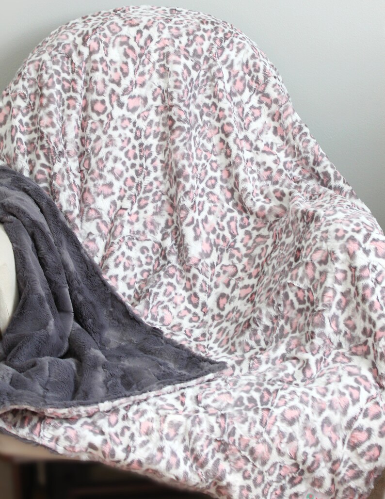 May include: A soft, plush throw blanket with a pink and gray leopard print on one side and a solid gray plush on the other side. The blanket is draped over a chair.