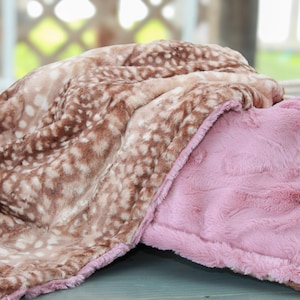 May include: A soft, plush blanket featuring a brown and white spotted pattern on one side and a solid light pink colour on the reverse. The fabric has a textured, fuzzy appearance with visible folds and layers, showcasing the two distinct sides.