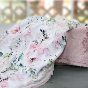 Puede incluir: Una manta suave con fondo blanco y un estampado floral de acuarela en tonos rosa, crema y verde salvia. El reverso de la manta est&aacute; hecho de una tela de felpa de color rosa claro, texturizada y s&oacute;lida, con un acabado suave y velloso.