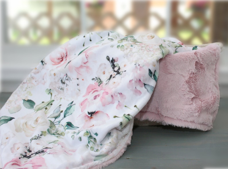 Puede incluir: Una manta suave con fondo blanco y un estampado floral de acuarela en tonos rosa, crema y verde salvia. El reverso de la manta est&aacute; hecho de una tela de felpa de color rosa claro, texturizada y s&oacute;lida, con un acabado suave y velloso.