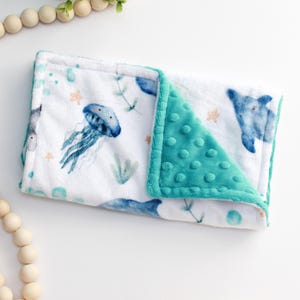 May include: A soft, white baby blanket with a teal border and backing. The blanket features a watercolor design of blue jellyfish, fish, and sea plants. The blanket is approximately 30 inches long and 20 inches wide.