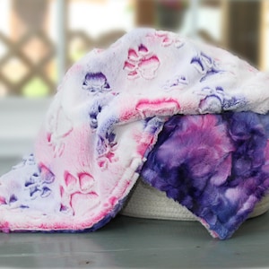 May include: A plush, soft-textured blanket featuring a white base with embossed paw prints in shades of pink and purple. The fabric displays a tie-dye effect with vibrant swirls of deep violet, magenta, and blue, creating a colourful, fuzzy surface.