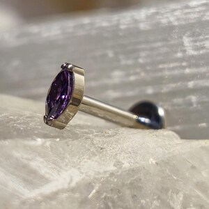 Pode incluir: Um piercing corporal prateado com uma pedra preciosa roxa escura com corte marquesa. A joia é colocada sobre um fundo branco texturizado, com um fundo desfocado.