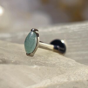 Könnte beinhalten: Ein silberner Ohrstecker mit einem hellblau-grünen, marquiseförmigen Edelstein. Der Ohrring hat einen schwarzen Rücken und ist auf einer strukturierten, weißen Oberfläche platziert.