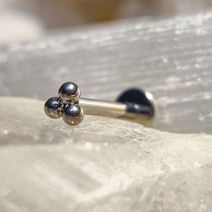 Op de afbeelding: Een zilverkleurige labret stud met drie kleine, ronde, zilveren kralen die bij elkaar gegroepeerd zijn. De sieraden worden gepresenteerd op een wit, kristallijn oppervlak, met een vage achtergrond van vergelijkbaar materiaal.