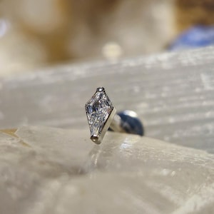 May include: A small, silver-colored piercing stud with a diamond-shaped, clear gemstone setting. The gemstone appears to be composed of multiple facets, reflecting light. The stud is set against a blurred, neutral background.
