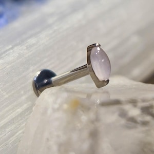 Könnte beinhalten: Ein silberner Labret-Stecker mit einem hellrosa, marquiseförmigen Edelstein. Der Schmuck ist vor einem Hintergrund aus weißen und blauen Kristallen platziert, was eine weiche, natürliche Ästhetik erzeugt. Der Stecker ist für Lippen- oder Ohrpiercings konzipiert.