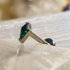 May include: A silver-colored earring with a marquise-cut, emerald-green gemstone. The earring has a flat back and a post for piercing. The gemstone is set in a silver bezel.