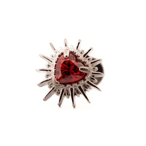 Puede incluir: Broche plateado con una gema roja oscura en forma de corazón en el centro. La gema está rodeada por un diseño de rayos de sol con puntas. El broche está sobre un fondo blanco.