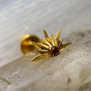 Puede incluir: Un pendiente de botón dorado con un diseño de sol y una pequeña gema roja redonda en el centro. El pendiente tiene un poste para la inserción y está colocado sobre un fondo claro y texturizado.