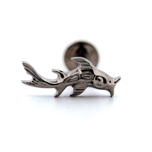 May include: A silver-coloured shark-shaped earring stud. The stud features a detailed design of a shark with fins and a rounded post for insertion. The earring is set against a white background.