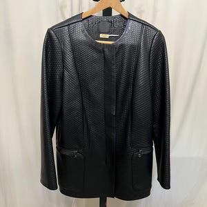 May include: A black jacket with a textured, quilted pattern. The jacket has a round neckline, long sleeves, and two front pockets with zippers. It is hanging on a wooden hanger on a black metal clothing rack.