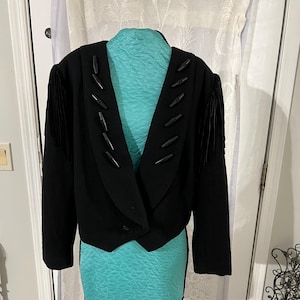 May include: A black cropped jacket with decorative wooden accents along the lapel and fringe detailing on the sleeves. The jacket is displayed on a turquoise dress form. The jacket has a single button closure.