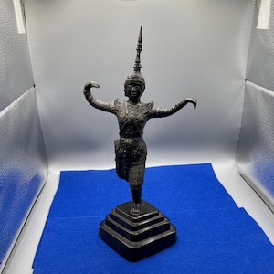 May include: A dark metallic statue of a figure in a traditional dance pose, featuring an ornate pointed headdress, detailed chest armor, and patterned clothing. The figure stands on a square, tiered base with arms extended outward and upward.